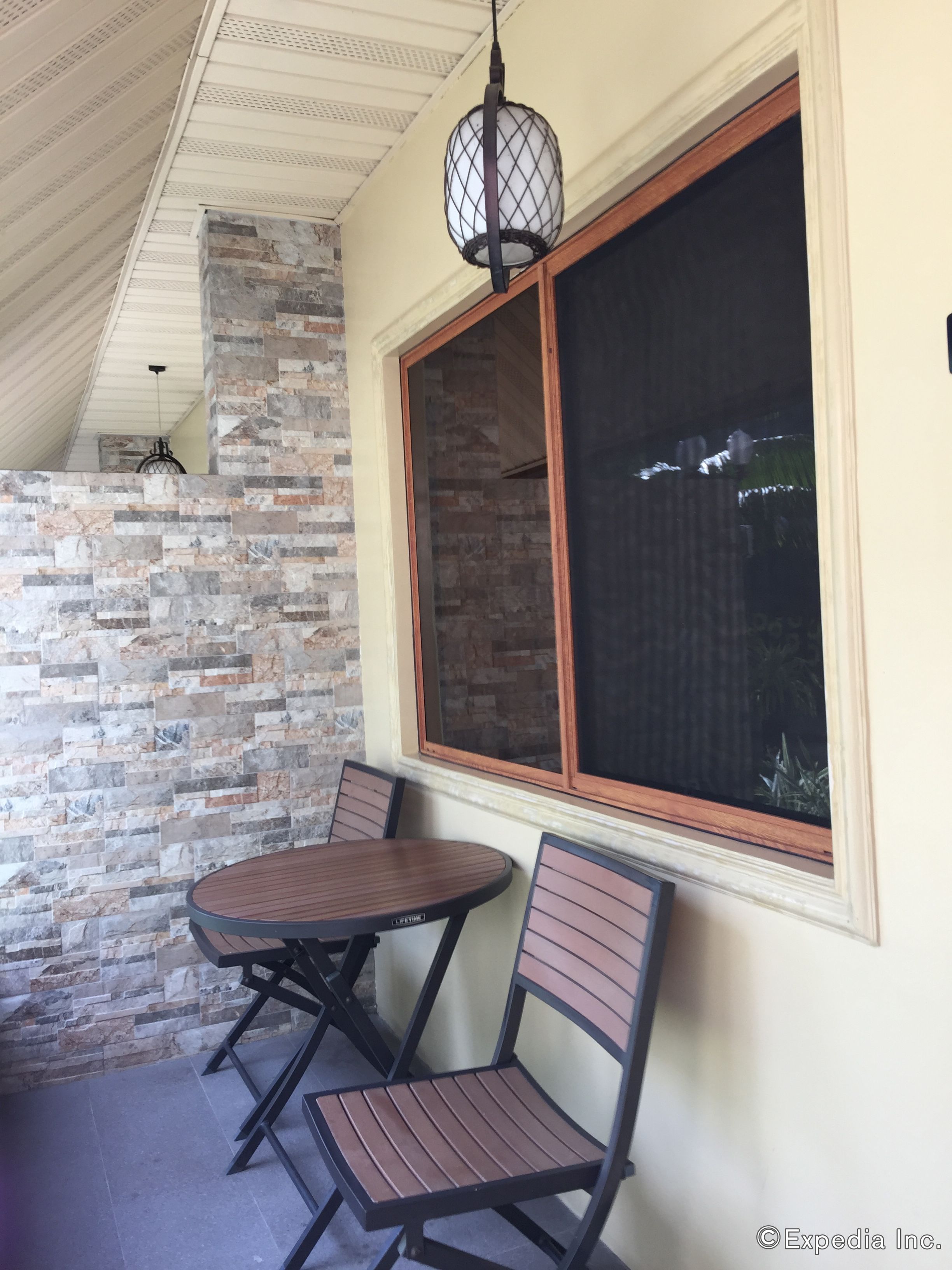 A patio area with a stone wall, a round wooden table, two chairs, and a large window with a view of greenery.