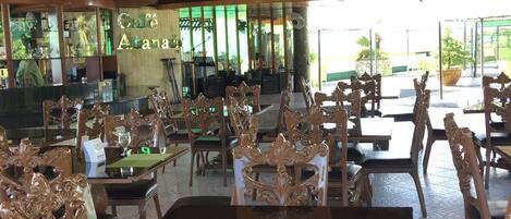 An indoor dining area with ornate wooden chairs and tables, a ceiling with exposed beams, and a view of the outside through large windows.
