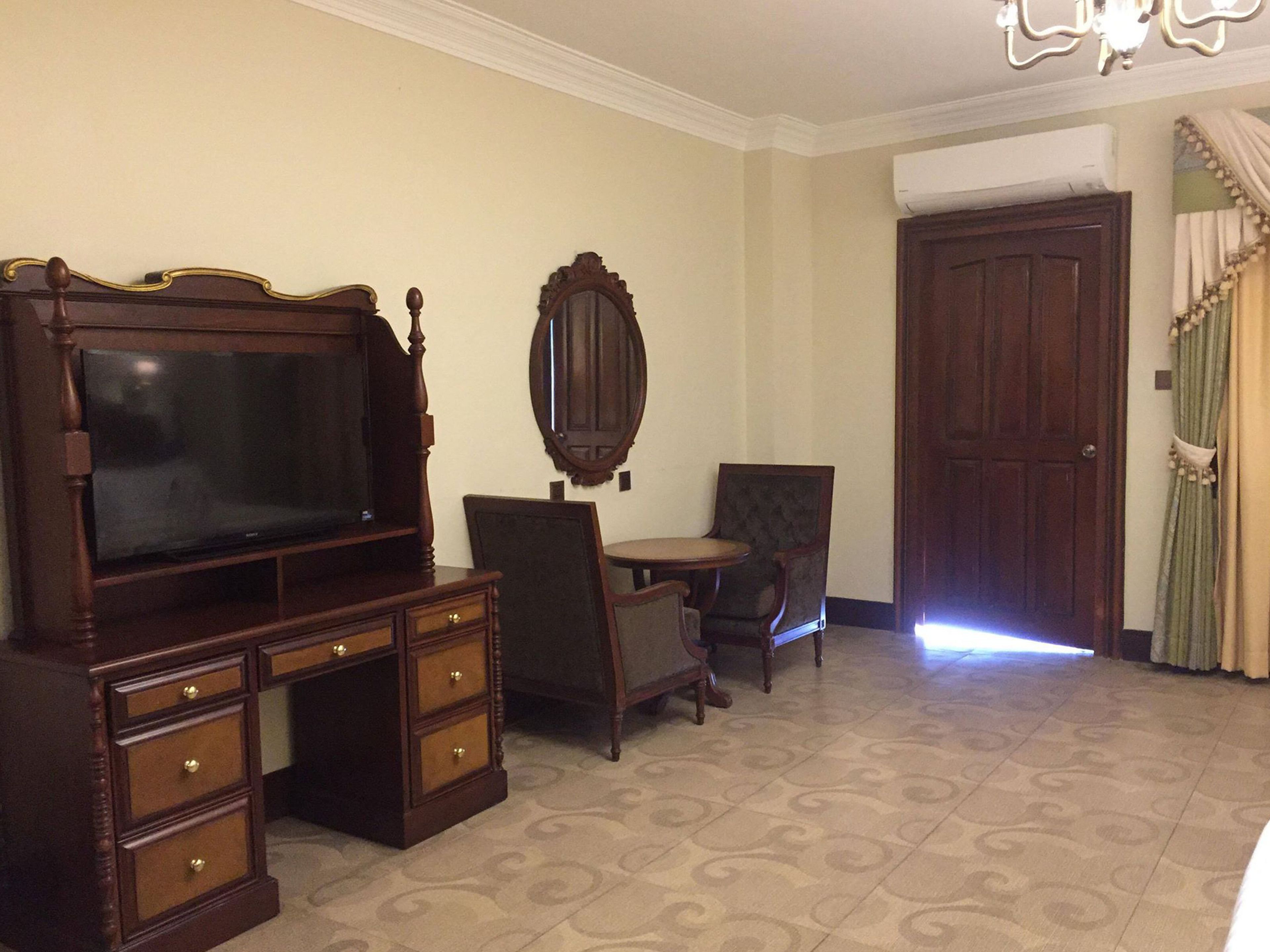 A room with a large wooden TV stand, a small round table, a chair, and a mirror on the wall.