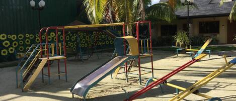 A playground with slides and swings, surrounded by palm trees and a building with a sloped roof.