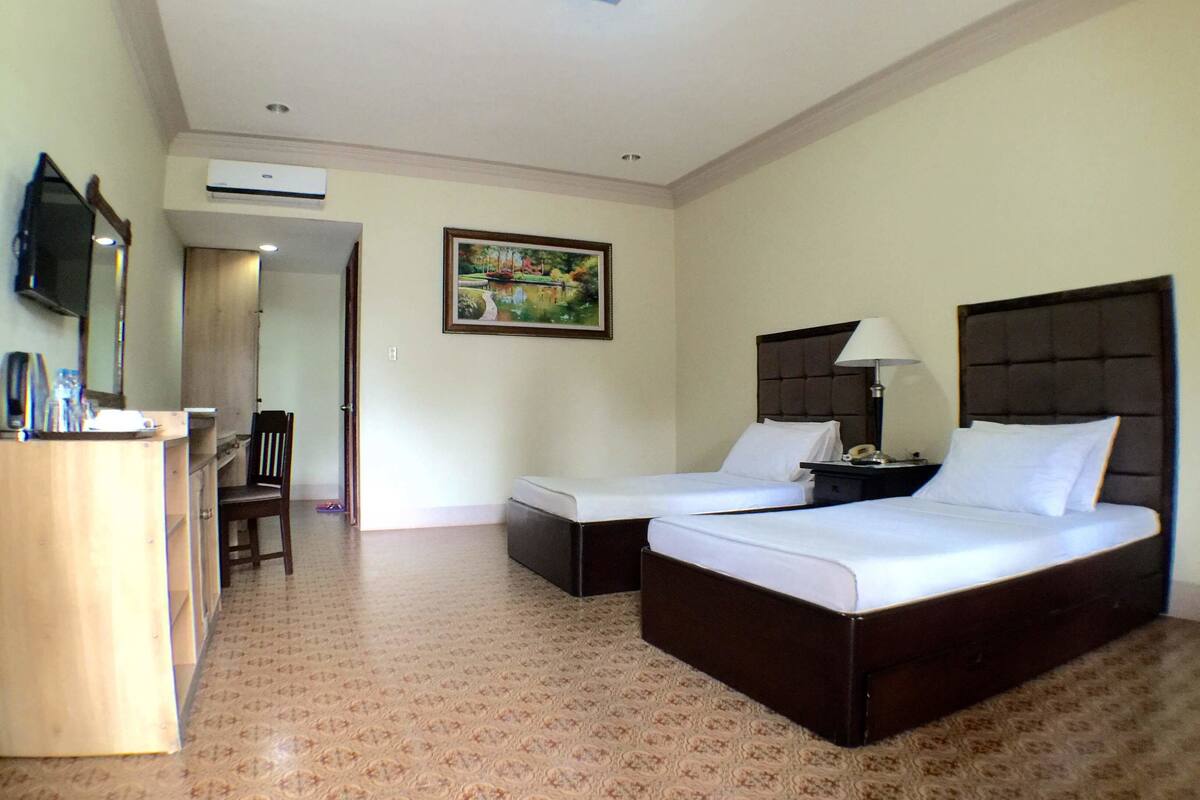A hotel room with two beds, a desk, a chair, a TV, and an air conditioning unit.