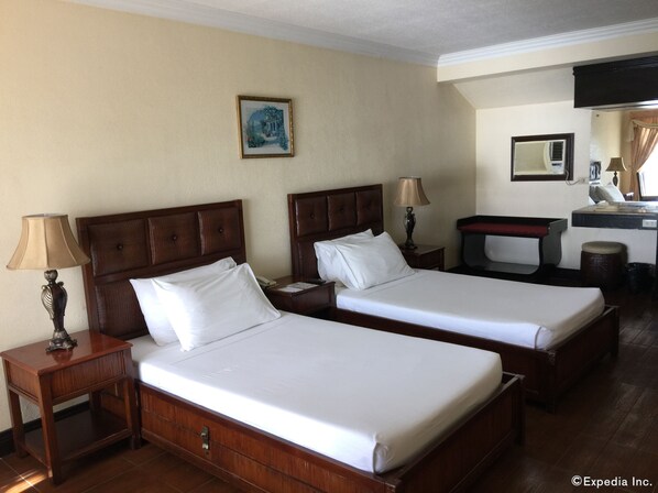 Una habitación de hotel con dos camas individuales, cabeceros de madera, mesitas de noche con lámparas, un escritorio pequeño con una silla y un cuadro en la pared.