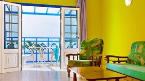 A room with a yellow wall, a wooden bench, a green sofa, and a table. There is a door leading to an outdoor area with a blue railing. - Atlantis Las Lomas (Tías)