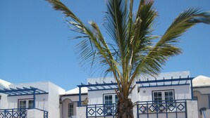 A building with white walls and blue balconies, surrounded by a landscaped garden with palm trees and shrubs. - Atlantis Las Lomas (Tías)