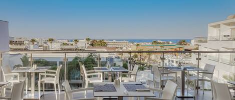Eine Dachterrasse mit weißen Tischen und Stühlen, die Blick auf die Stadt und das Meer bietet.