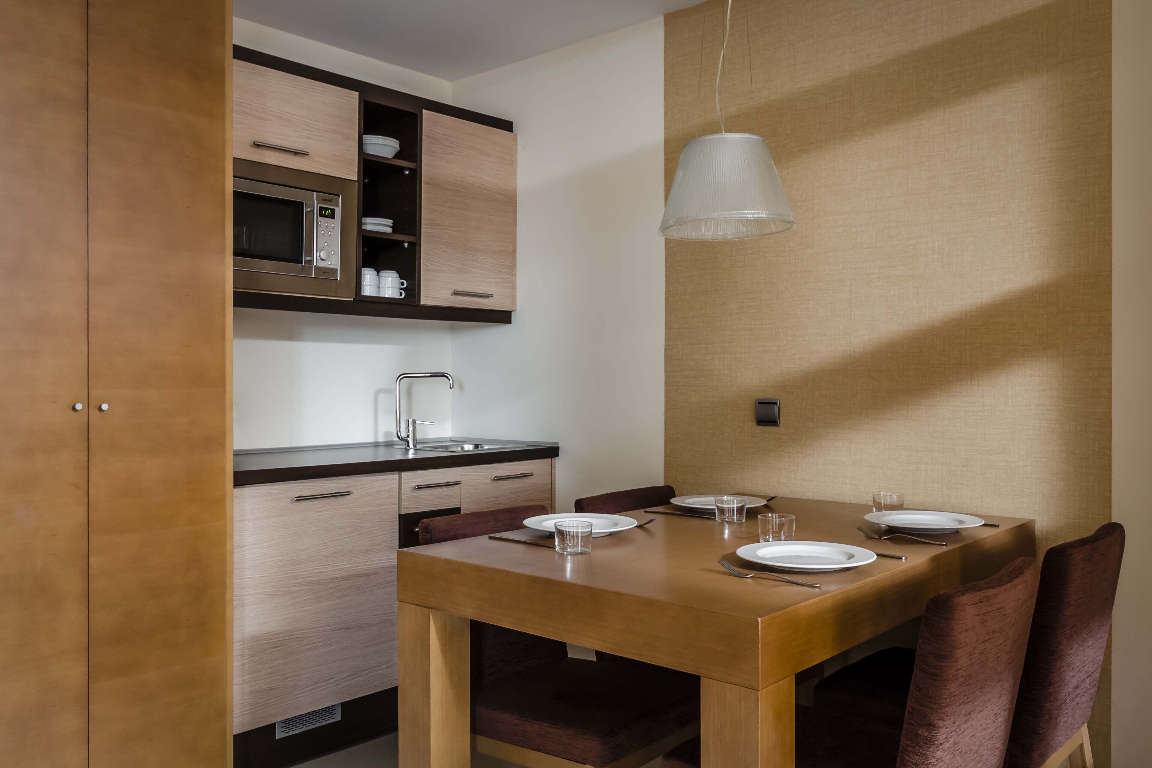 A compact kitchen with a microwave, wooden cabinets, and a dining table set for two with chairs.