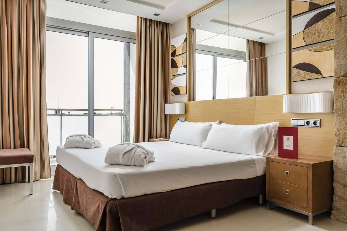A hotel room with a large bed, a bedside table with a lamp, a red folder, and a view of the city through the window.