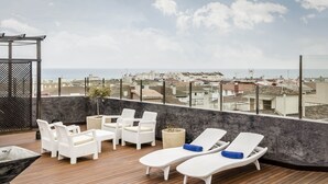 Een terras op het dak met witte rieten meubels, een vuurkuil en uitzicht op de stad en de zee.