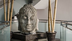 Een stenen Boeddhabeeld hoofd op een voetstuk in een moderne binnenruimte met glazen hekwerken en bamboeplanten.
