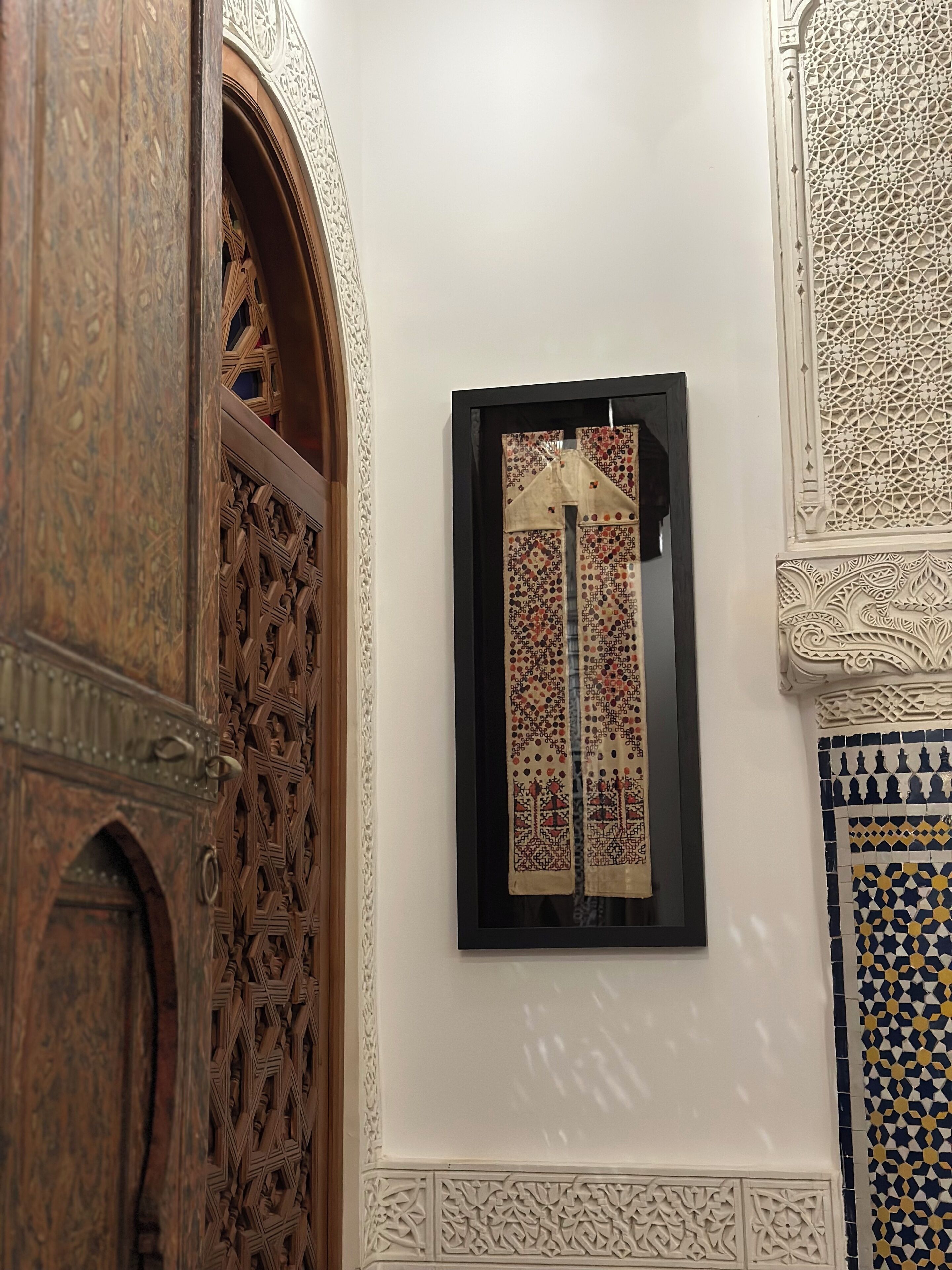 Um quarto com porta de madeira com esculturas detalhadas, tapeçaria emoldurada com padrões geométricos e parede com mosaico azul e amarelo.