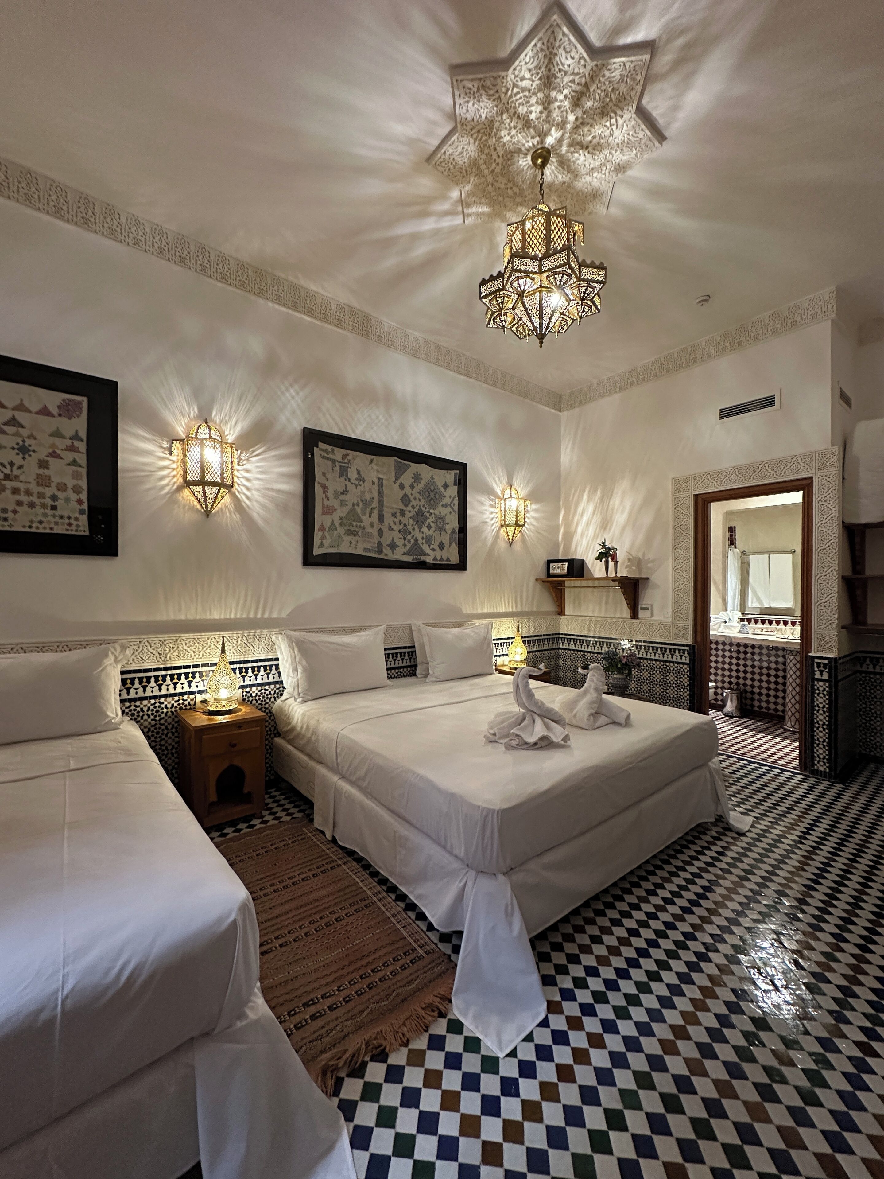 A hotel room with two beds, a chandelier, wall sconces, a framed tapestry, and a checkered floor.
