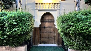 Una puerta de madera tradicional con detalles de metalurgia elaborada, bordeada por arbustos verdes exuberantes, que da paso a un vestíbulo con baldosas.