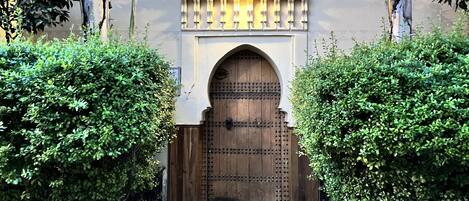 A traditional wooden door with intricate metalwork, flanked by lush green bushes, leading to a tiled entrance.