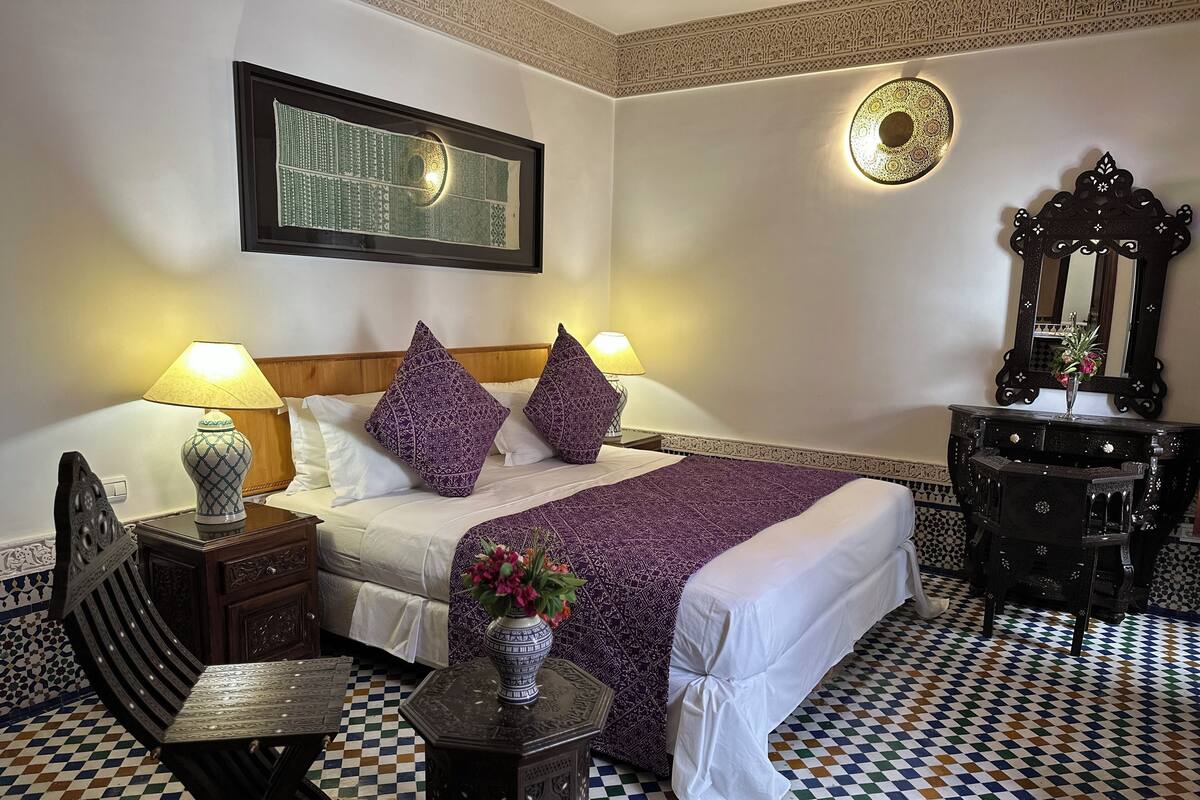 A hotel room with a checkered floor, a bed with patterned pillows, a nightstand with a lamp, a decorative mirror, and a wall-mounted artwork.