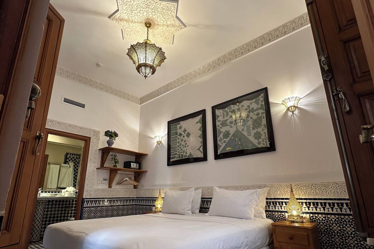 A bedroom with a large bed, a nightstand, a wall-mounted lamp, and a patterned rug.