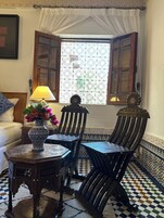 Quarto tradicional marroquino com piso xadrez, mesa estampada, cadeira de madeira e janela com vista.