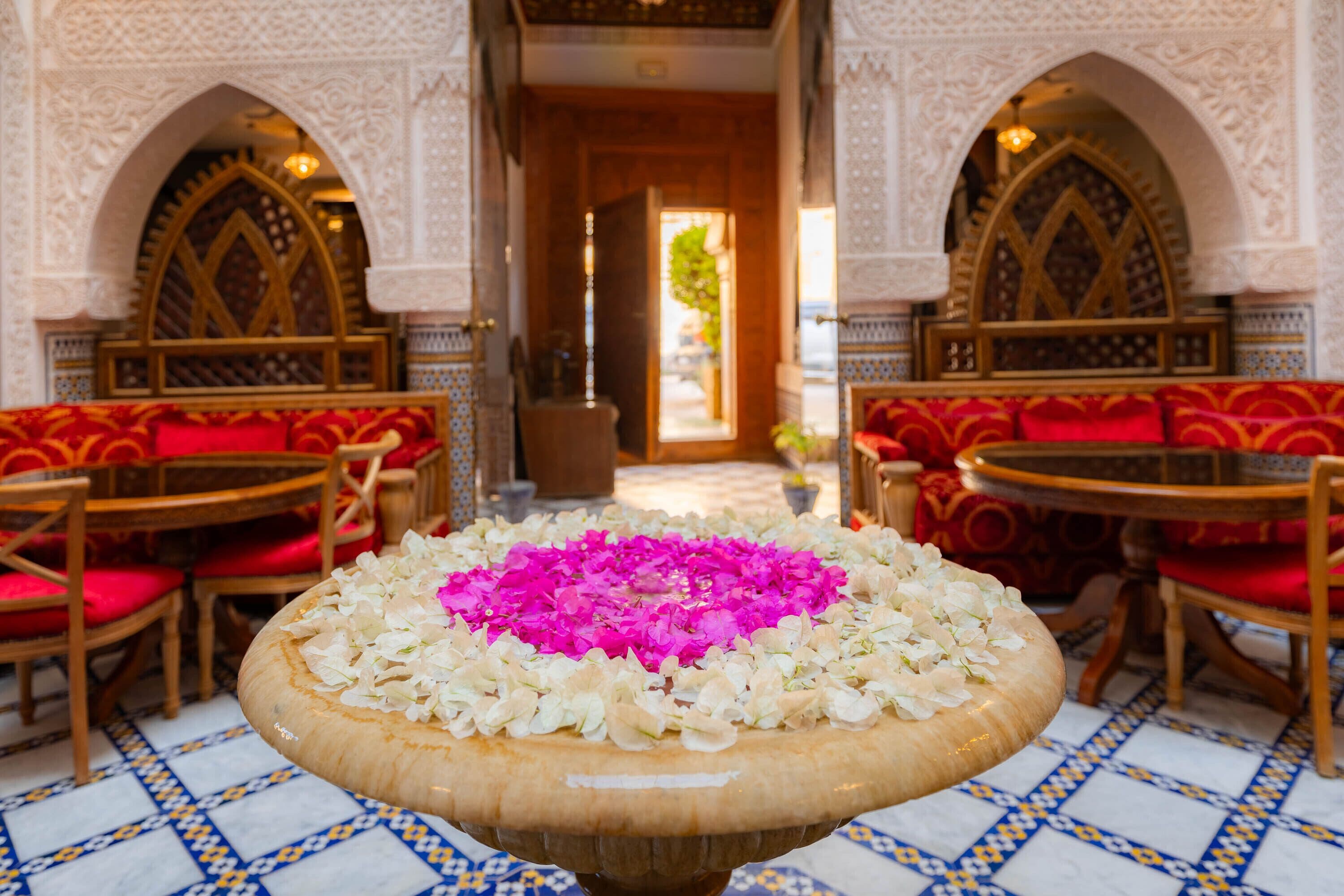 A room with a central table adorned with white and pink flowers, surrounded by red upholstered chairs and a patterned floor.
