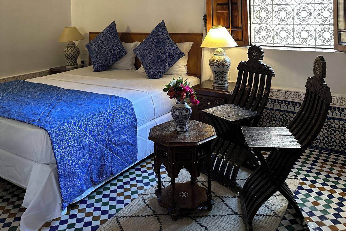 A hotel room with a bed, a table, two chairs, a decorative lamp, a window with a patterned screen, and a ceiling fan.