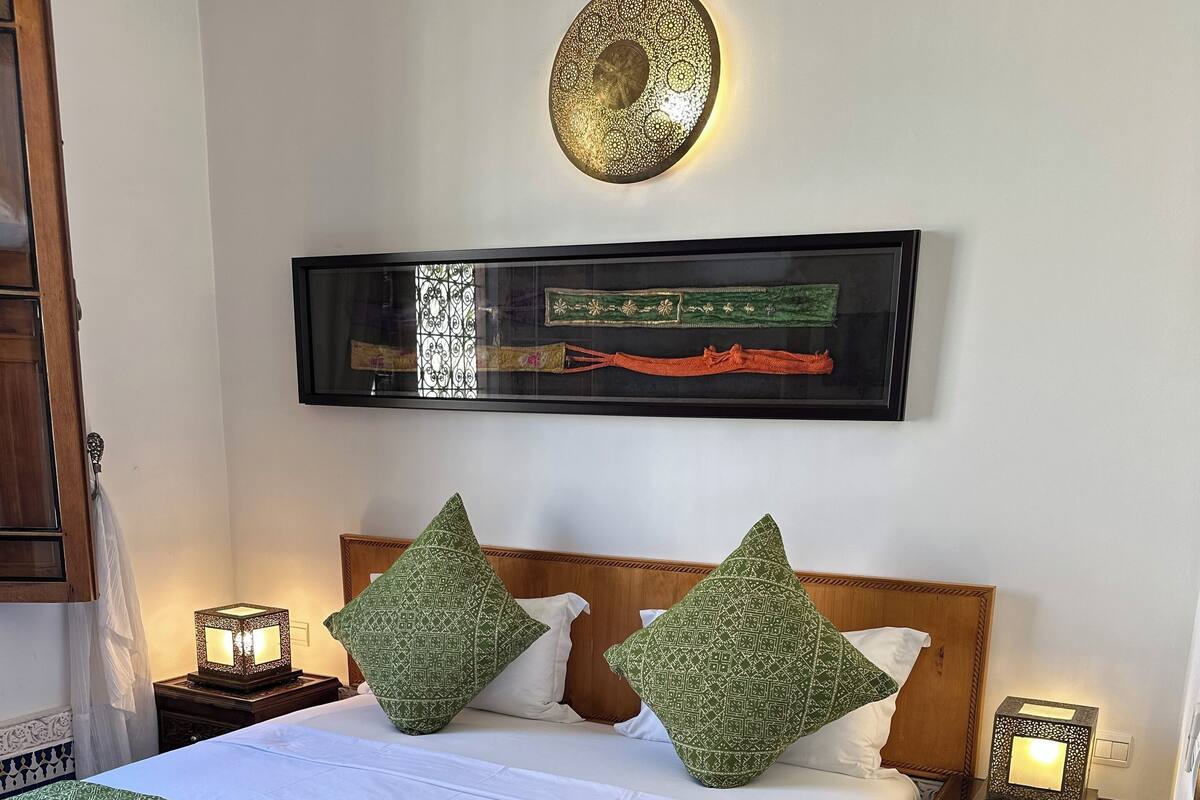 A neatly made bed with a green patterned bedspread, two pillows, and a headboard. There is a wall-mounted decorative plate above the bed, a small bedside table with a lamp, and a patterned rug on the floor.