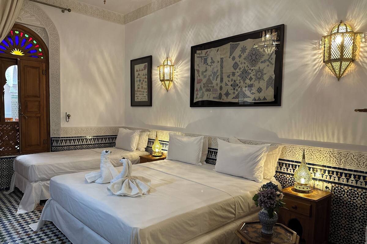 A hotel room with two beds, decorative wall sconces, a patterned tapestry, and a checkered floor.
