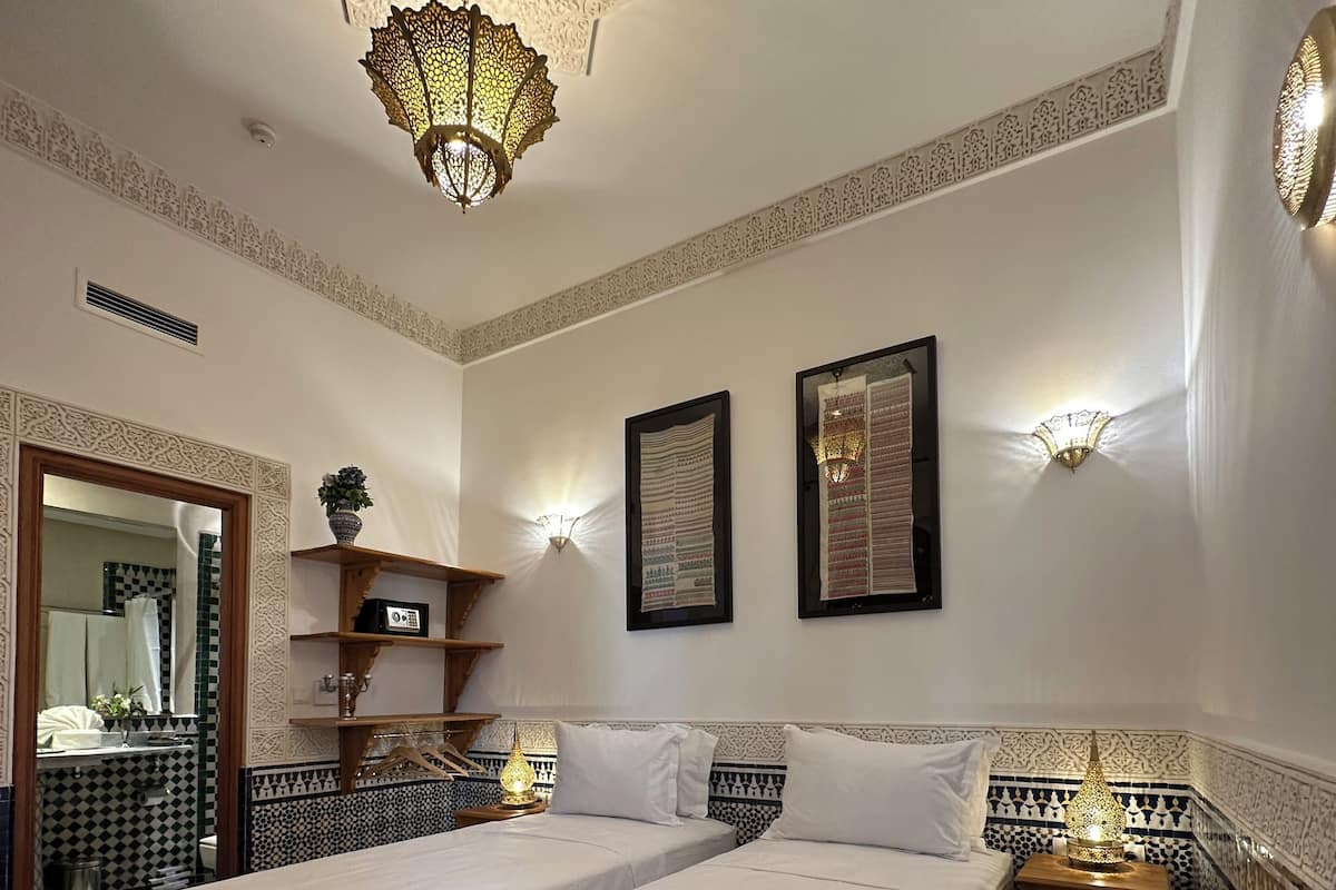 A hotel room with two beds, a decorative ceiling light, wall sconces, a mirror, and patterned tiles.