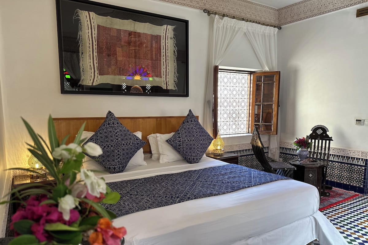 A neatly arranged bedroom with a bed, pillows, a nightstand, a lamp, a window with curtains, a chair, and a decorative rug.