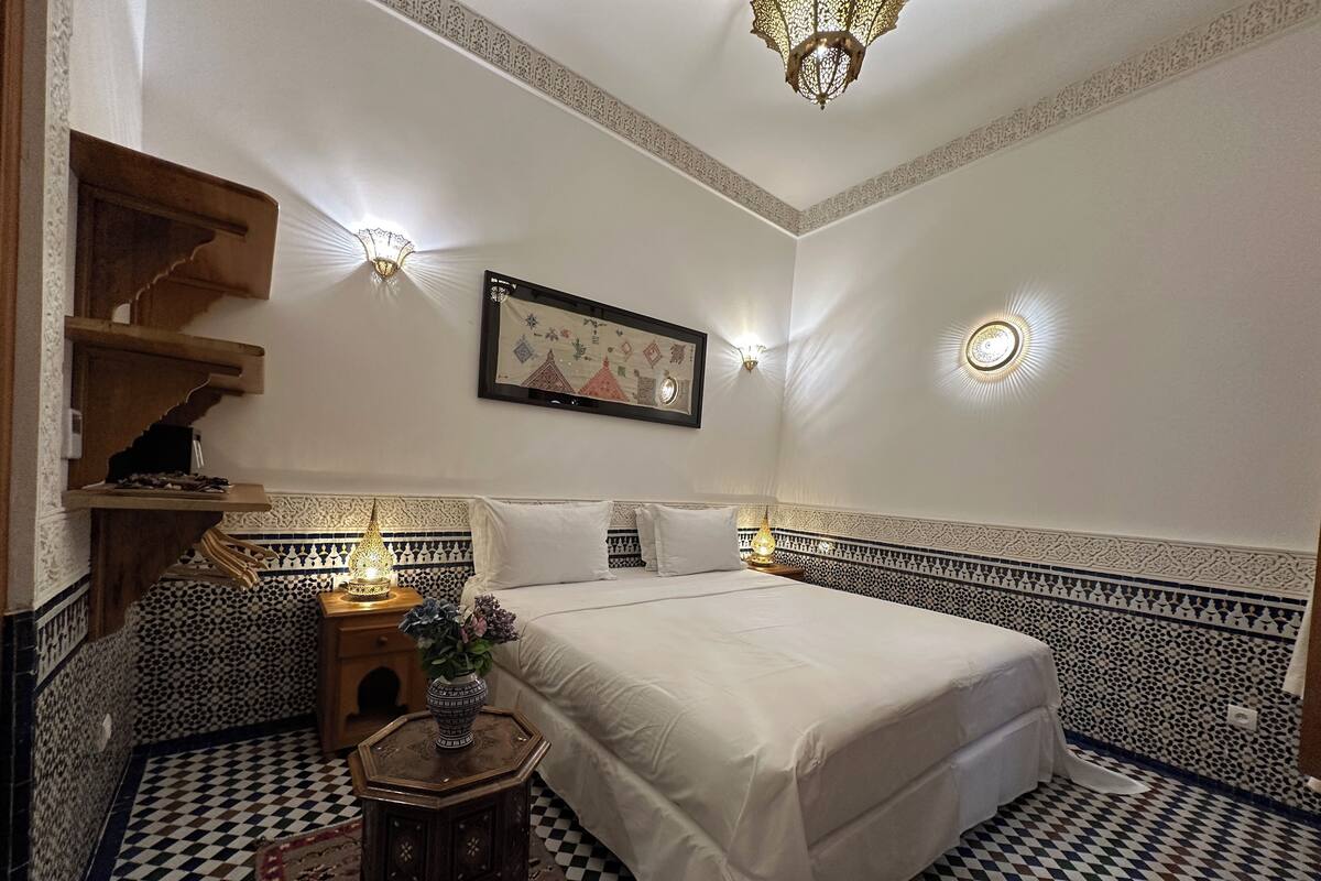 A bedroom with a large bed, decorative wall sconces, a patterned rug, and a small side table with a vase.