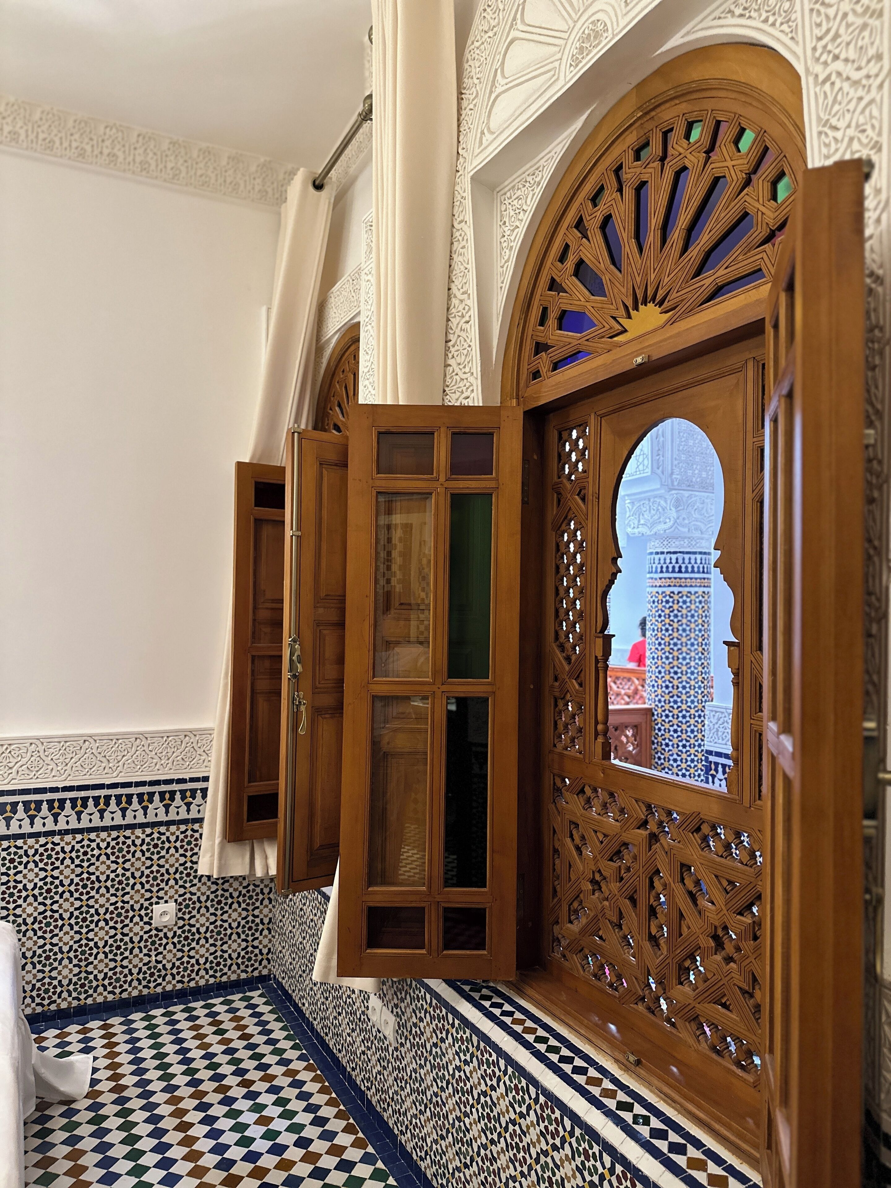 A room with intricate tile work, a wooden door with glass panels, and a view of a building through an arched window.