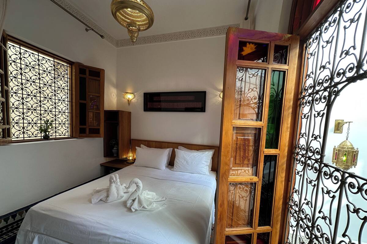 A neatly made bed with white linens and two towels shaped like swans, a wooden headboard, a wall-mounted light, and a decorative iron gate.