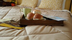 A neatly made bed with a quilted cover, two pillows with a yellow and white pattern, a green mug, and a tray with two rolls of bread and some papers. - Casa Mariella (Naples)