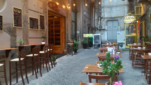 An outdoor café with wooden tables and chairs, potted plants, and chalkboard menus. - Casa Mariella (Naples)