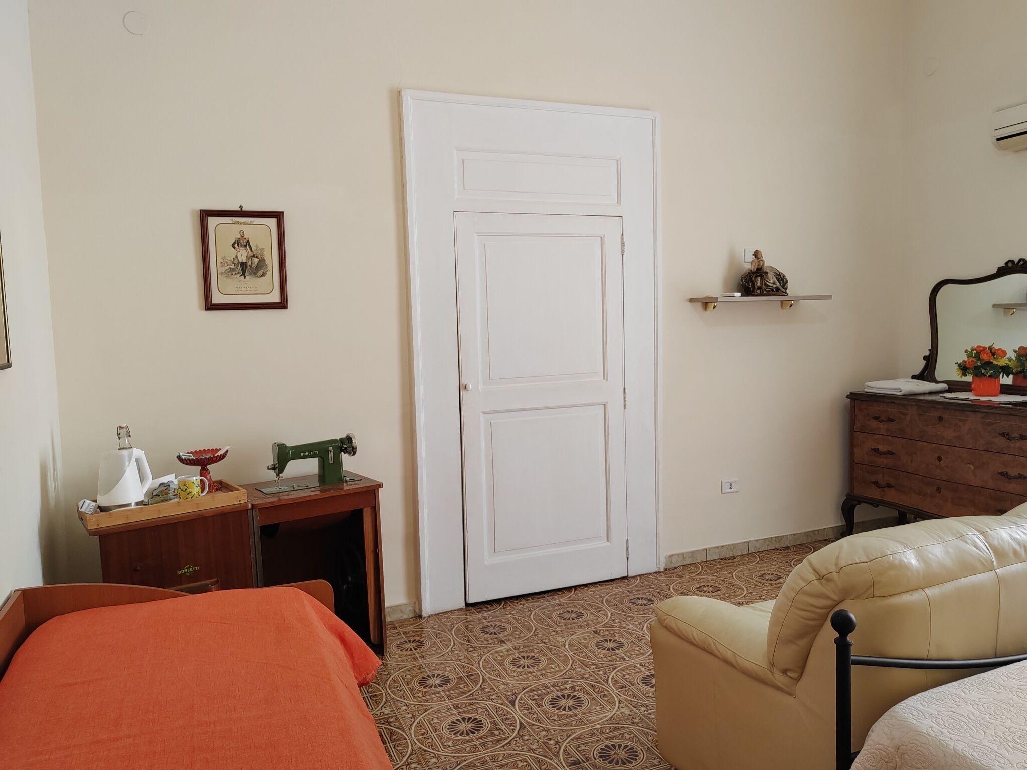 Una stanza con un letto, una piccola scrivania con una macchina da cucire, una cassettiera, uno specchio e un quadro appeso al muro.