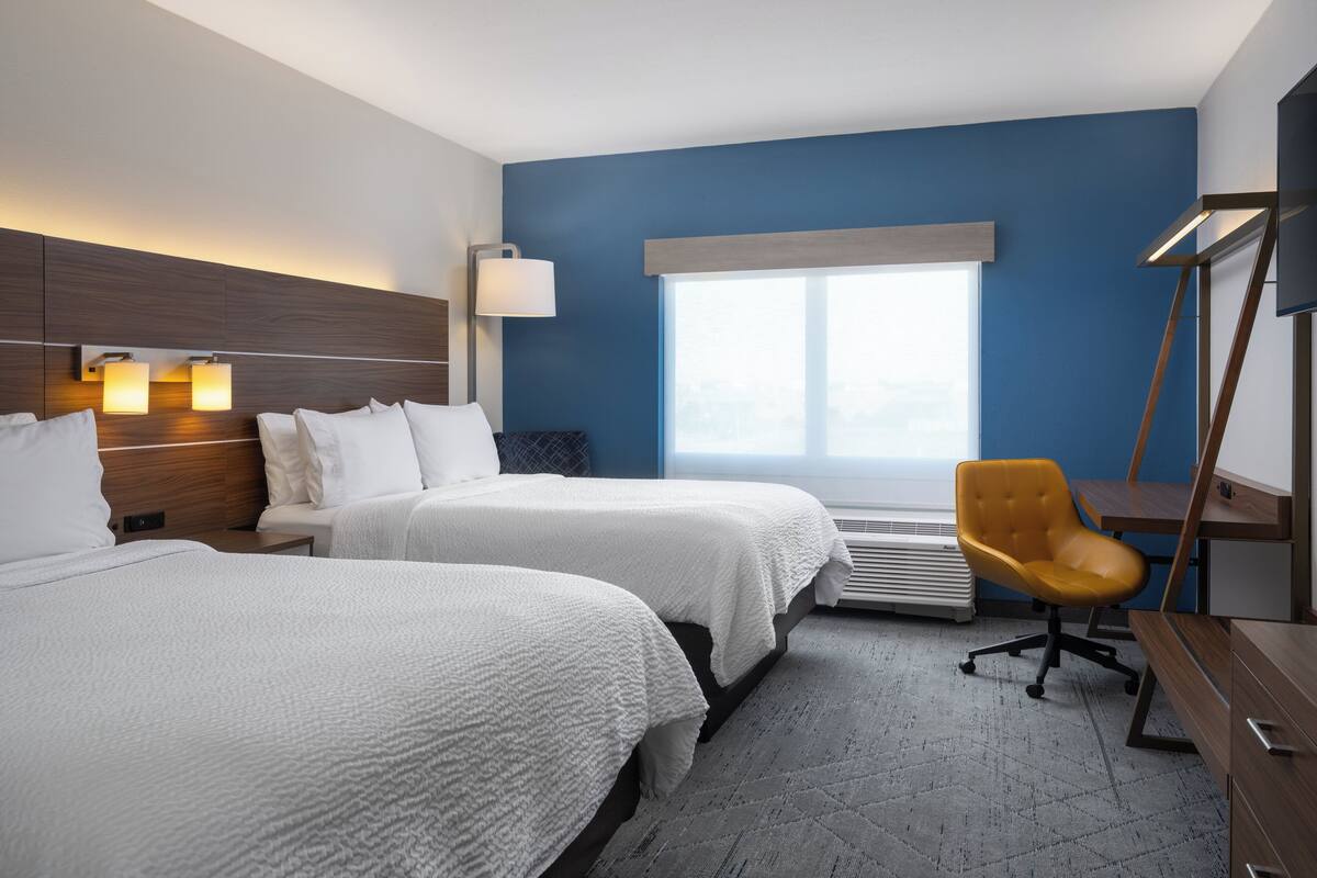 A hotel room with two beds, a desk, a chair, a large window, and a wall-mounted TV.