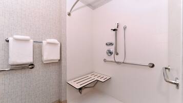 Un baño con paredes de azulejos blancos, una ducha con barra de apoyo y dos toallas blancas en un perchero.