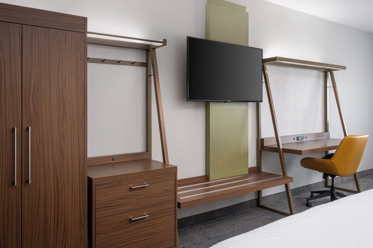 A bedroom with a wooden wardrobe, a desk with a chair, and a mounted TV.