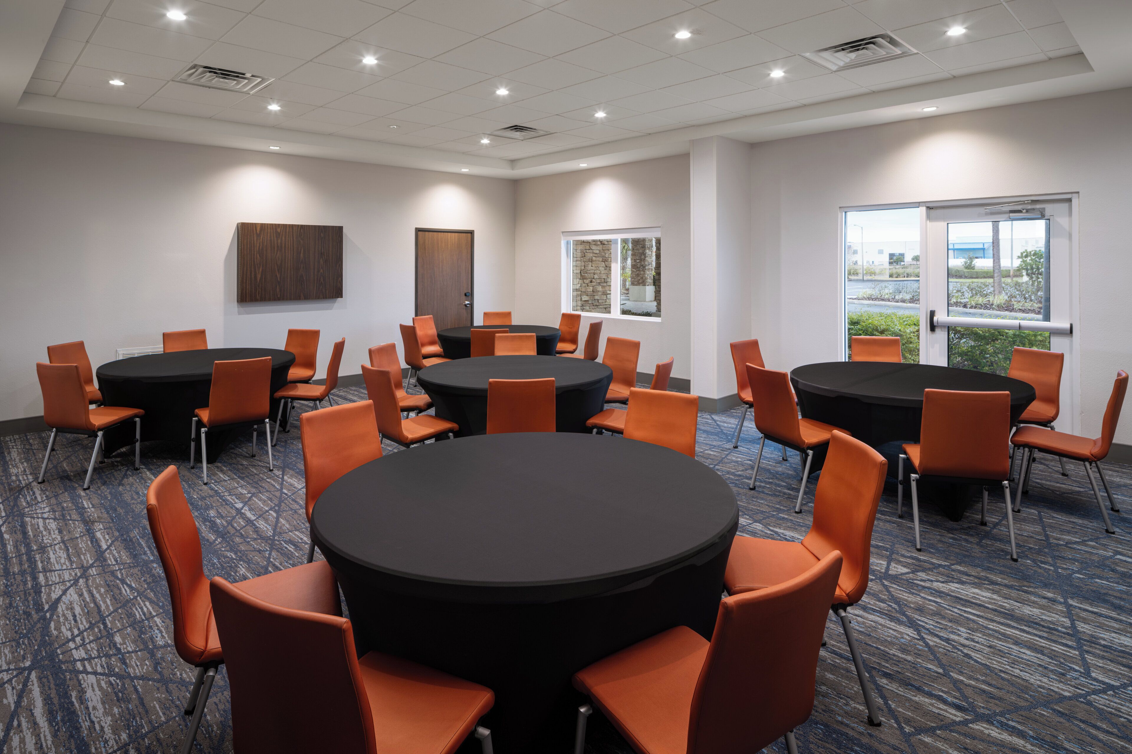 A conference room with multiple tables and orange chairs, large windows, and a view of the outdoors.