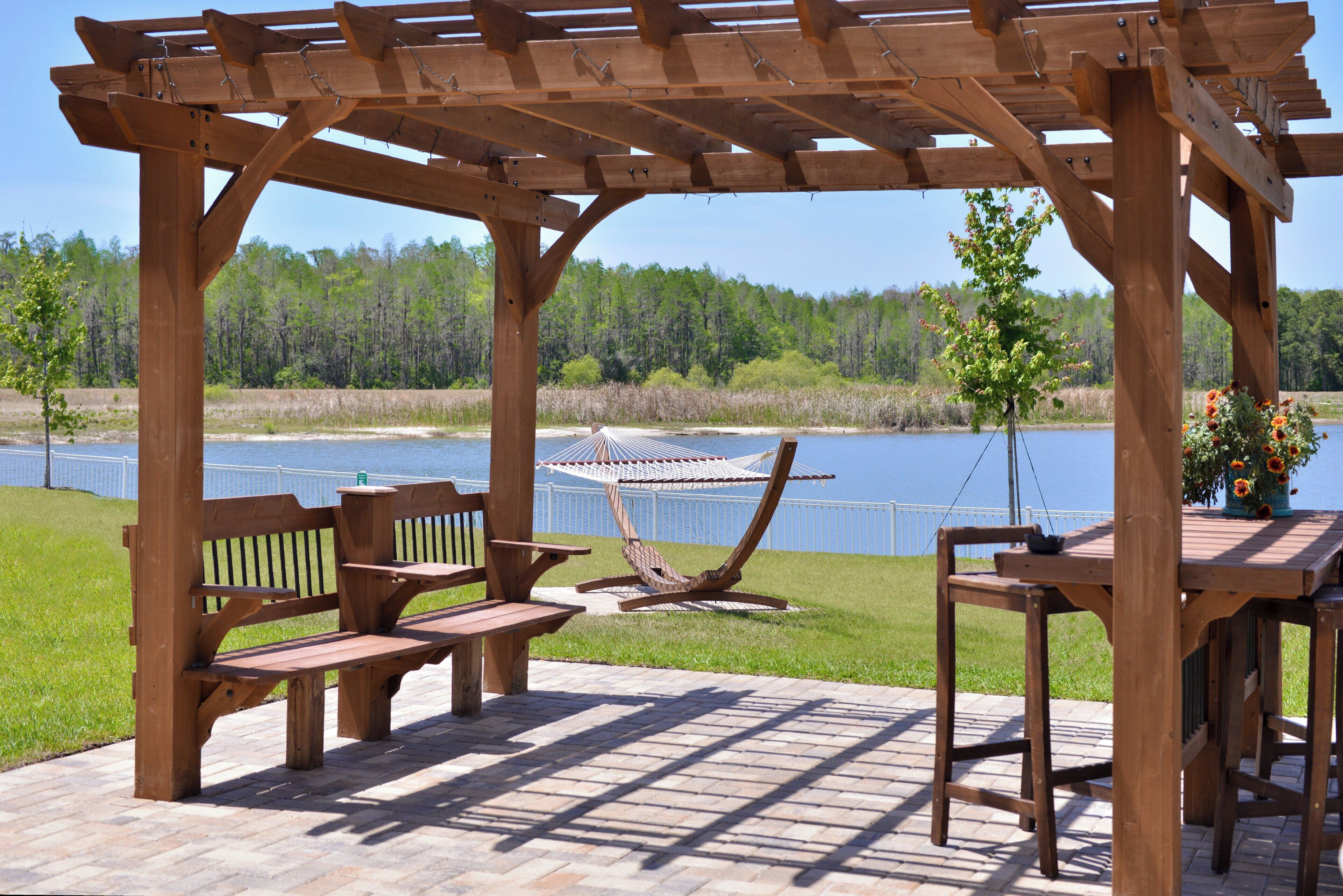 A wooden pergola with a bench and a table, overlooking a body of water and a forested area.