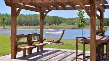 A wooden pergola with a bench and a table, overlooking a body of water and a forested area.