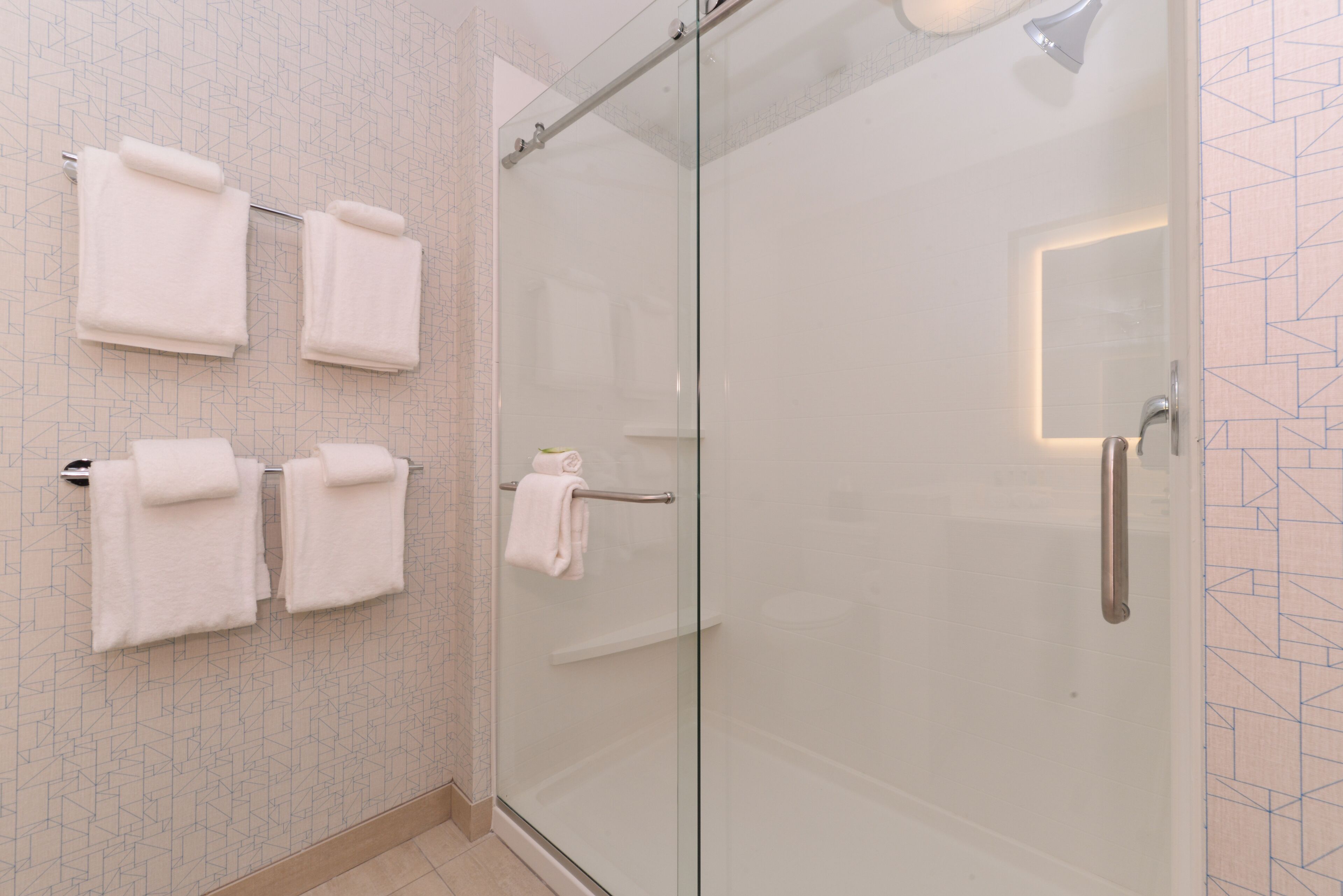 Un baño moderno con cabina de ducha de vidrio, múltiples toallas blancas en un perchero y un revestimiento de pared con estampado geométrico.