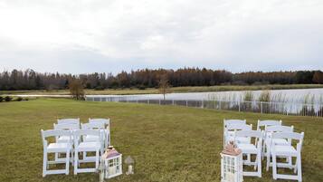 Un entorno exterior con filas de sillas blancas dispuestas para un evento, un cuerpo de agua y una zona boscosa al fondo.