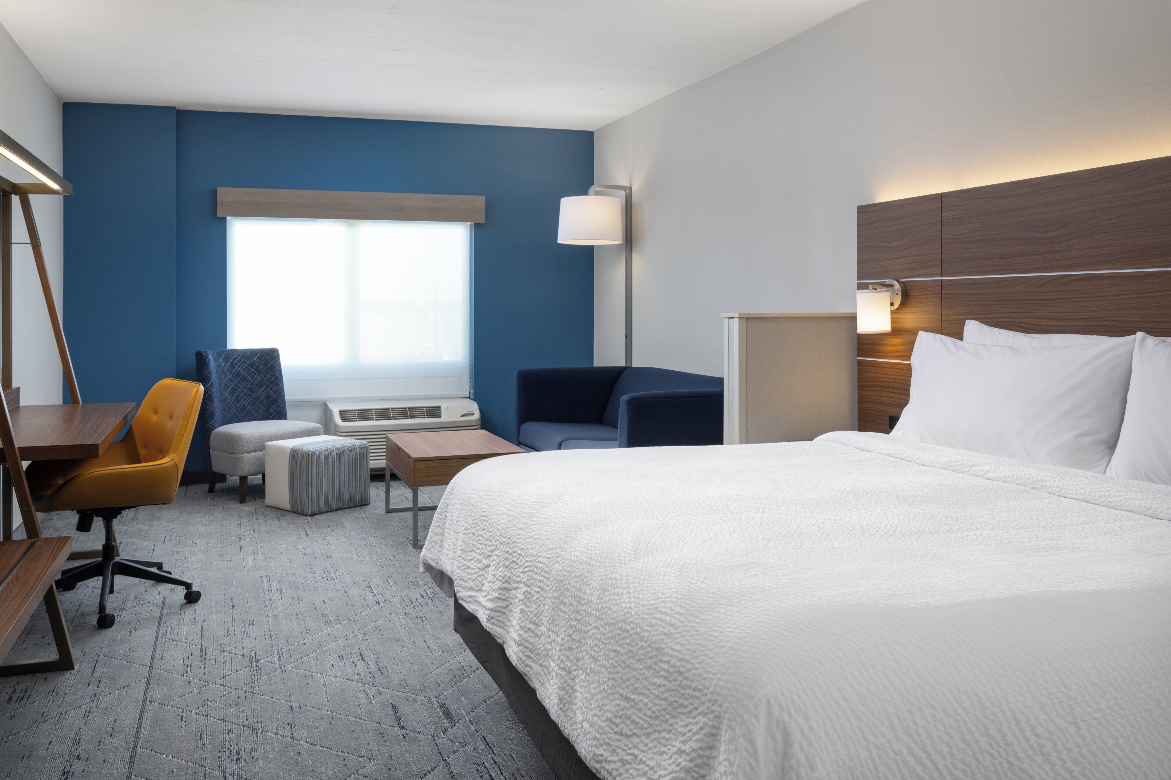 Habitación de hotel con una cama grande, un escritorio con silla, un sofá, una lámpara de pie y una ventana con persianas.