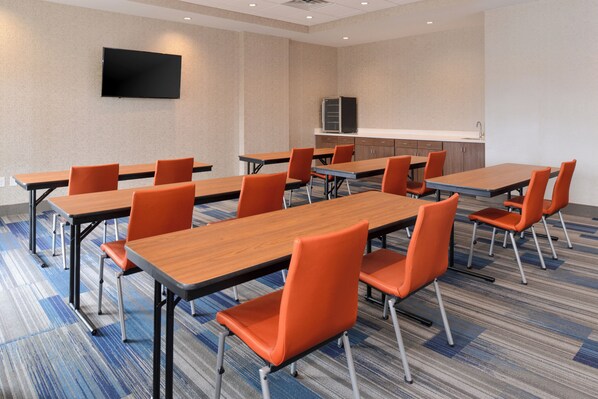 A conference room with multiple tables and chairs, a flat-screen TV mounted on the wall, and a kitchenette area.