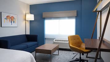 Habitación de hotel con una pared azul, una cama blanca, un escritorio de madera, una silla amarilla, un sofá azul y una ventana con persianas.
