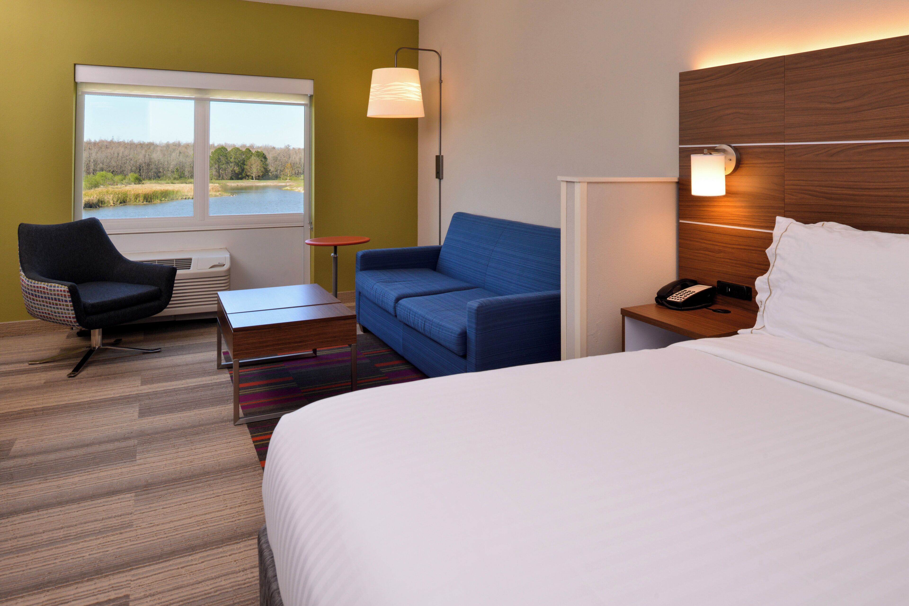 A hotel room with a bed, a blue sofa, a chair, a small table, a lamp, and a window with a view of a lake.