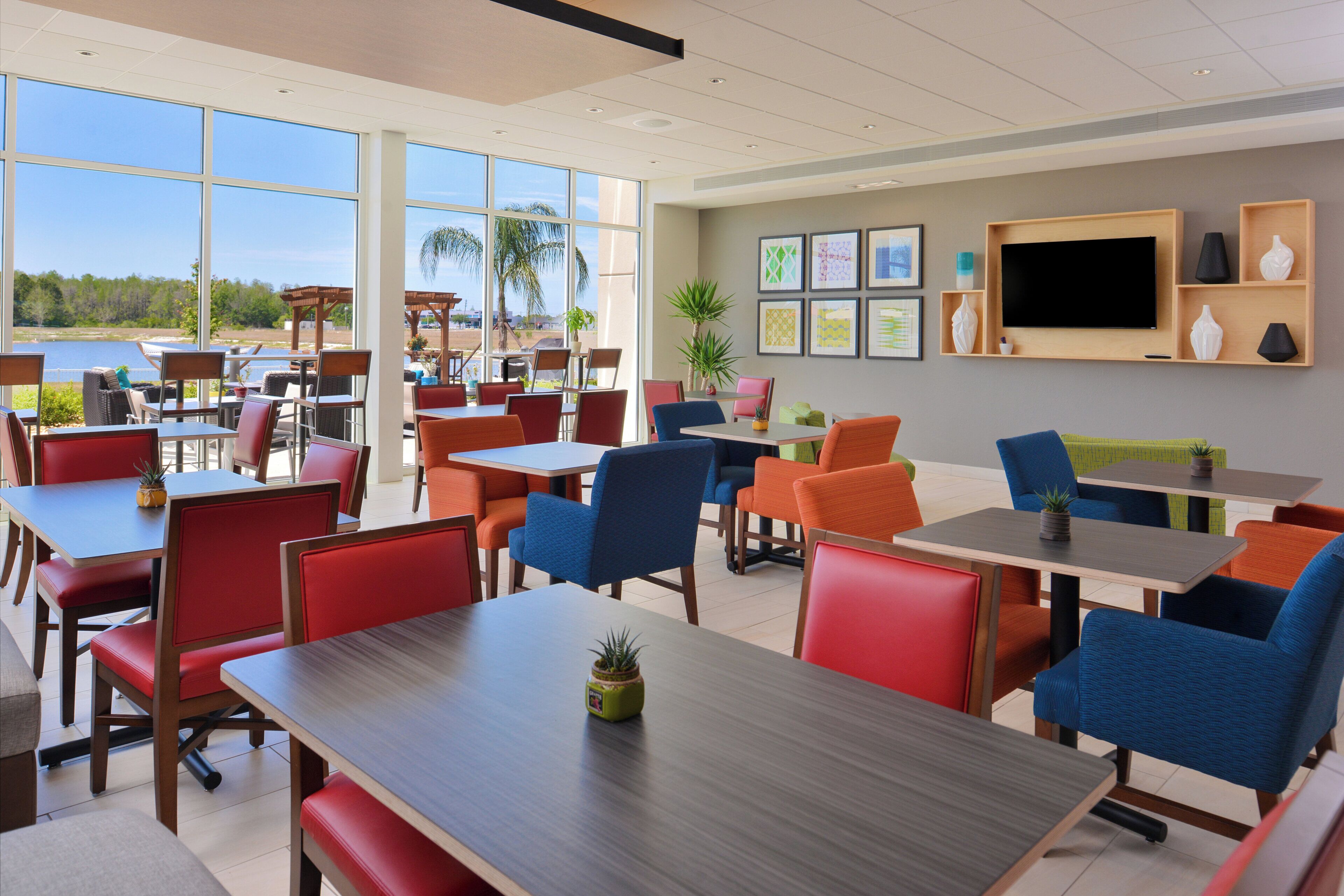 Un espace repas moderne avec plusieurs tables et chaises, de grandes fenêtres donnant sur l’extérieur et un téléviseur à écran plat fixé au mur.
