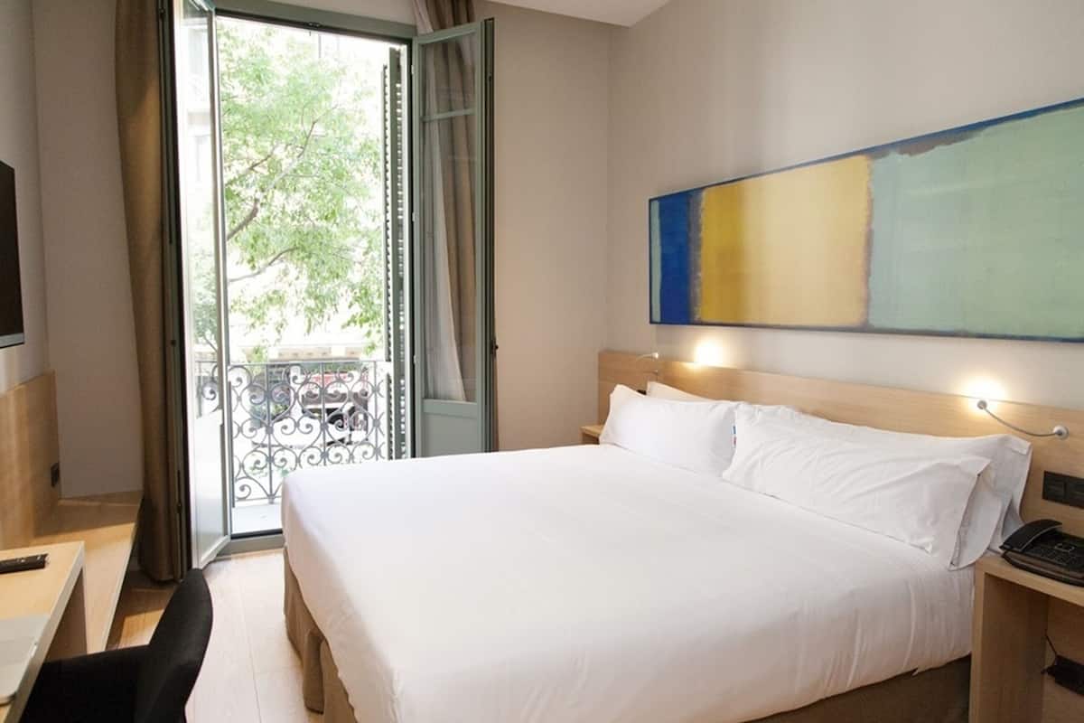 Habitación de hotel con cama, escritorio, silla, balcón con vista a árboles y un cuadro en la pared.
