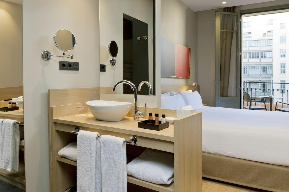 Habitación de hotel con una cama grande, lavamanos doble y balcón con vista a la ciudad.