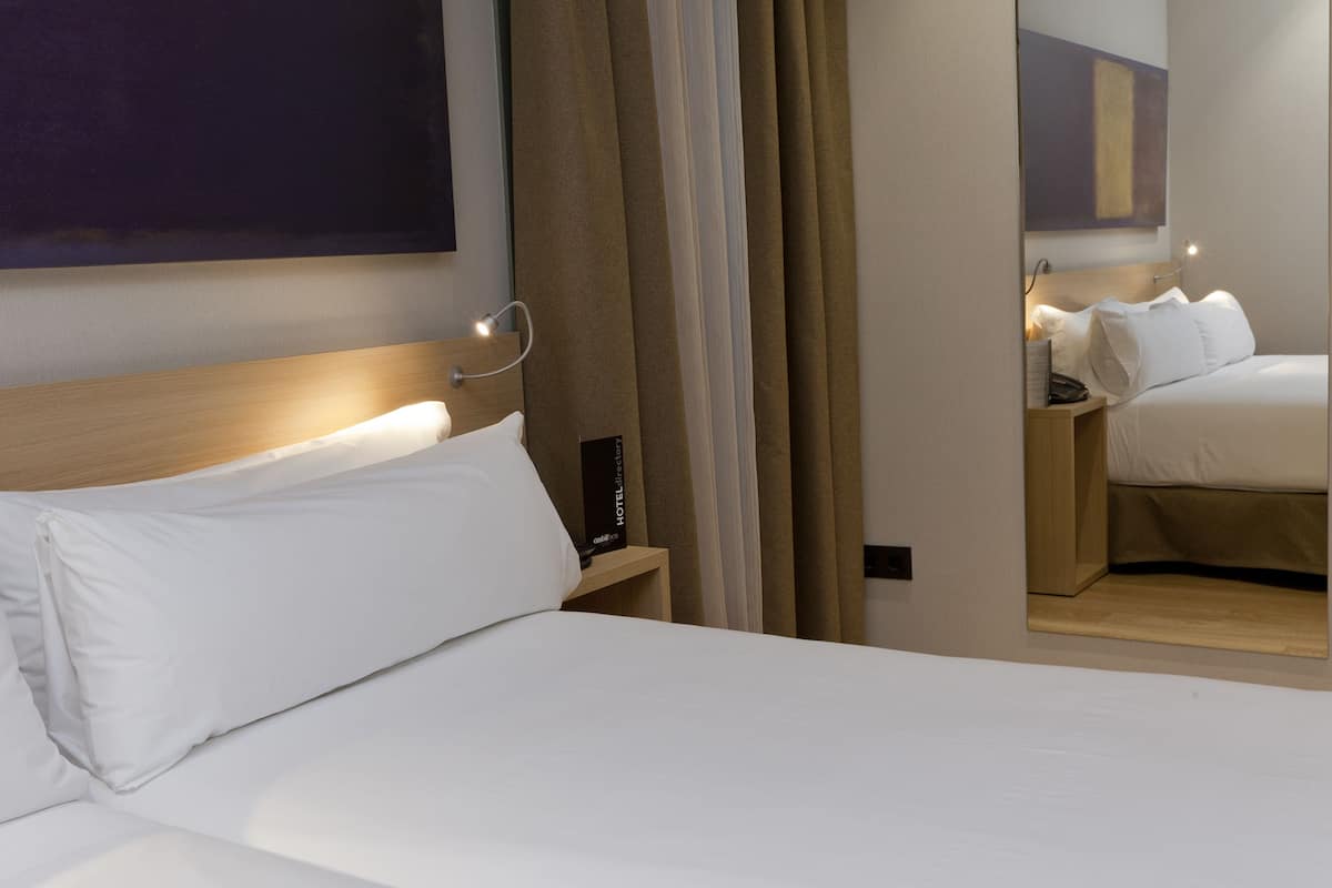 Habitación de hotel con una cama grande, una mesita de noche con un teléfono y un espejo que refleja la habitación.