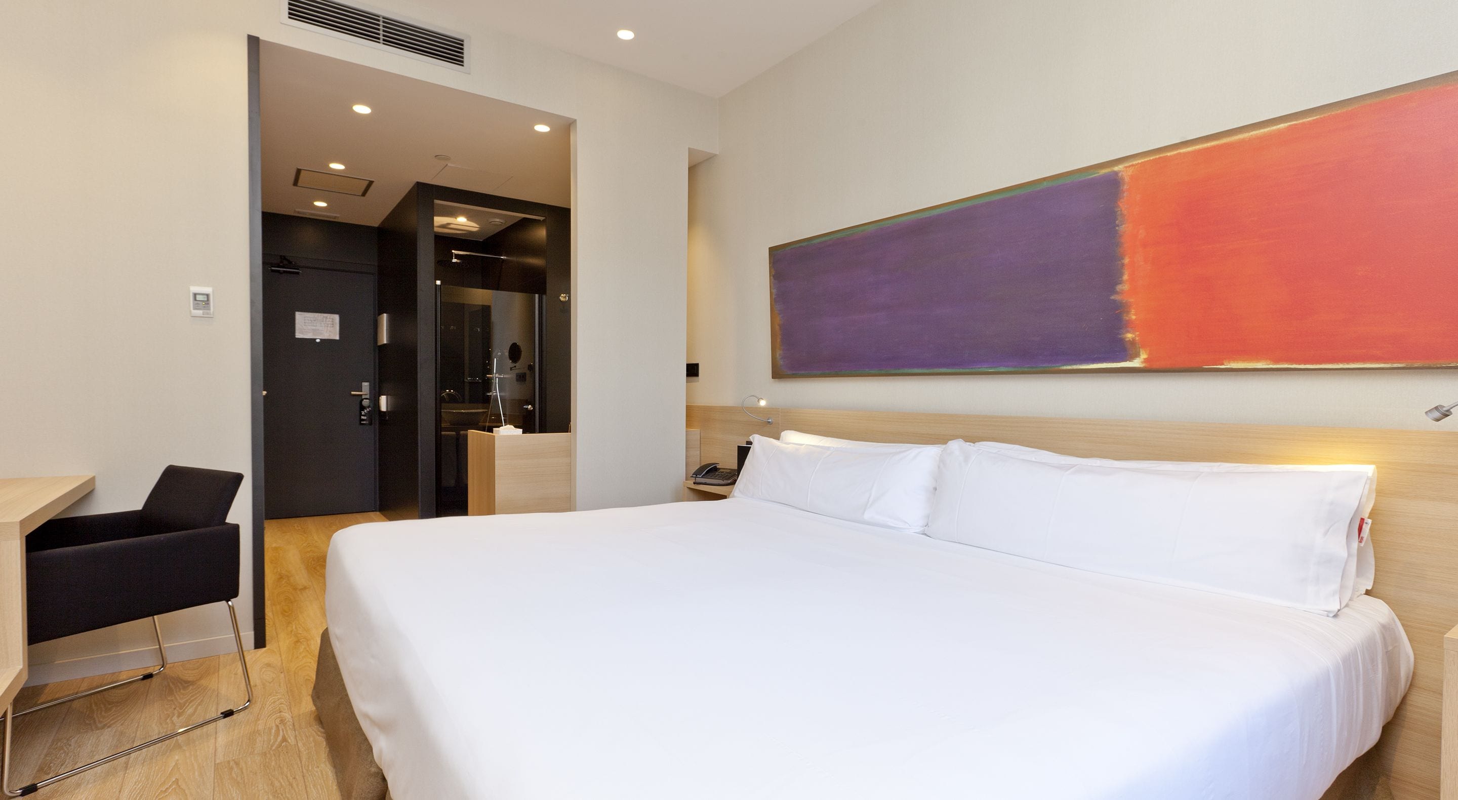 A hotel room with a large bed, a desk, and a chair. There is a painting on the wall and a door leading to a bathroom.