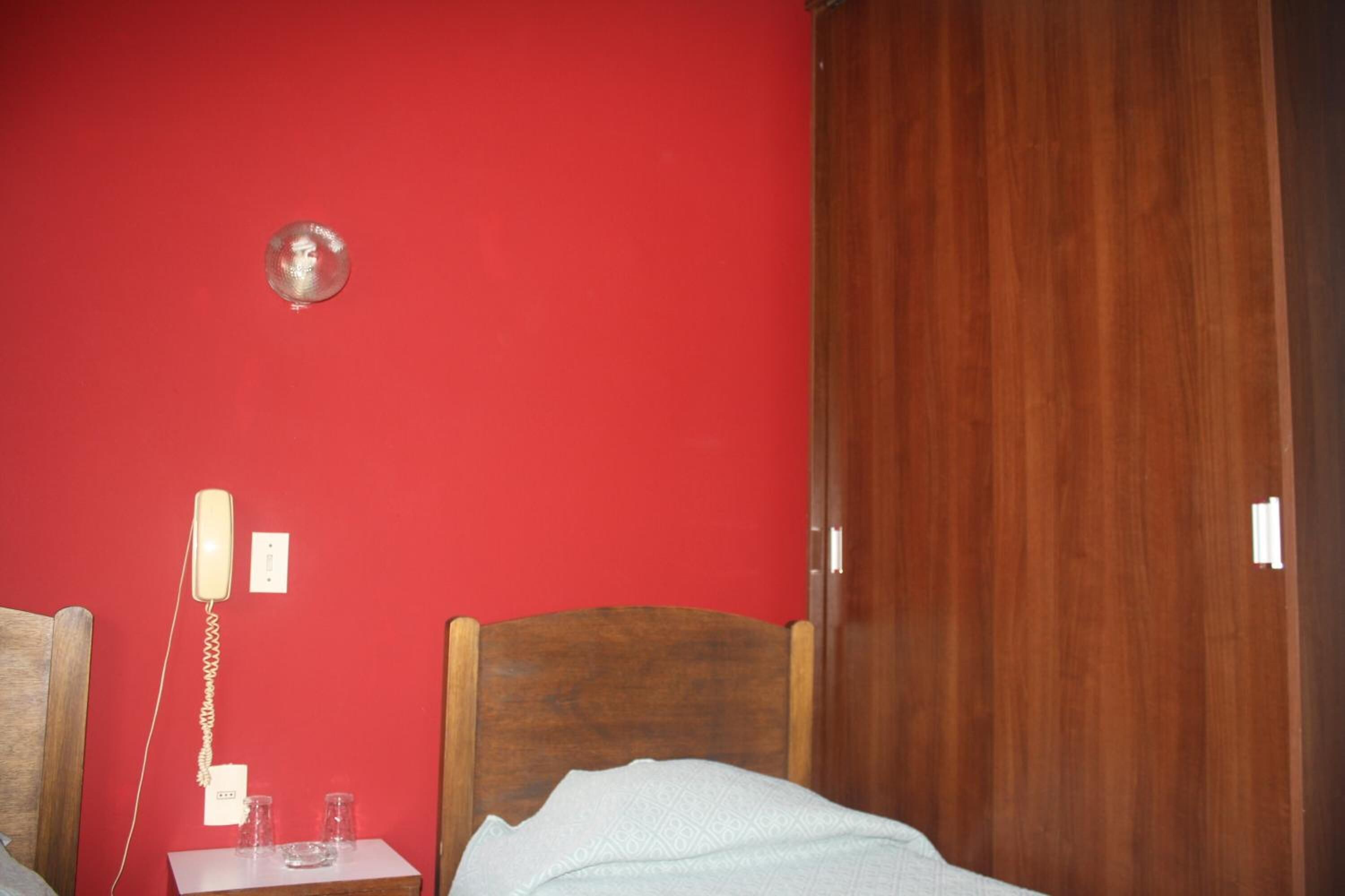Et værelse med en seng med trægavle, en rød væg, en telefon og et lille bord med glas og en kop.